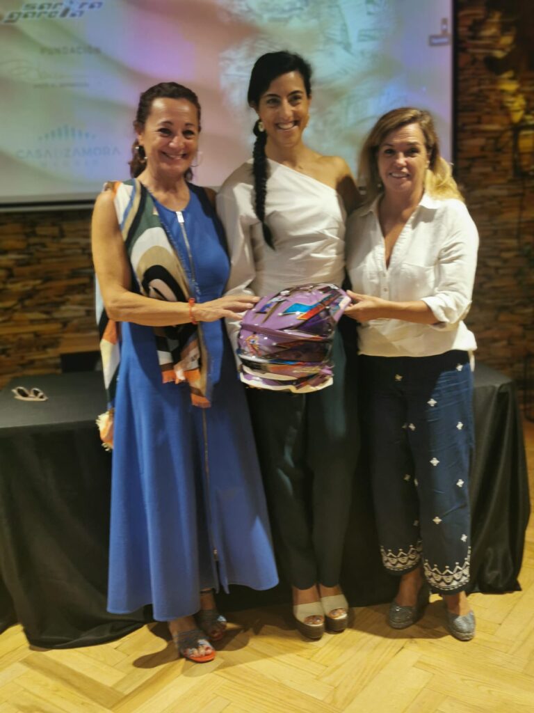 Sara García en el centro, sonriente, sosteniendo su casco decorado junto a dos representantes de la Fundación Blanca durante un acto oficial.